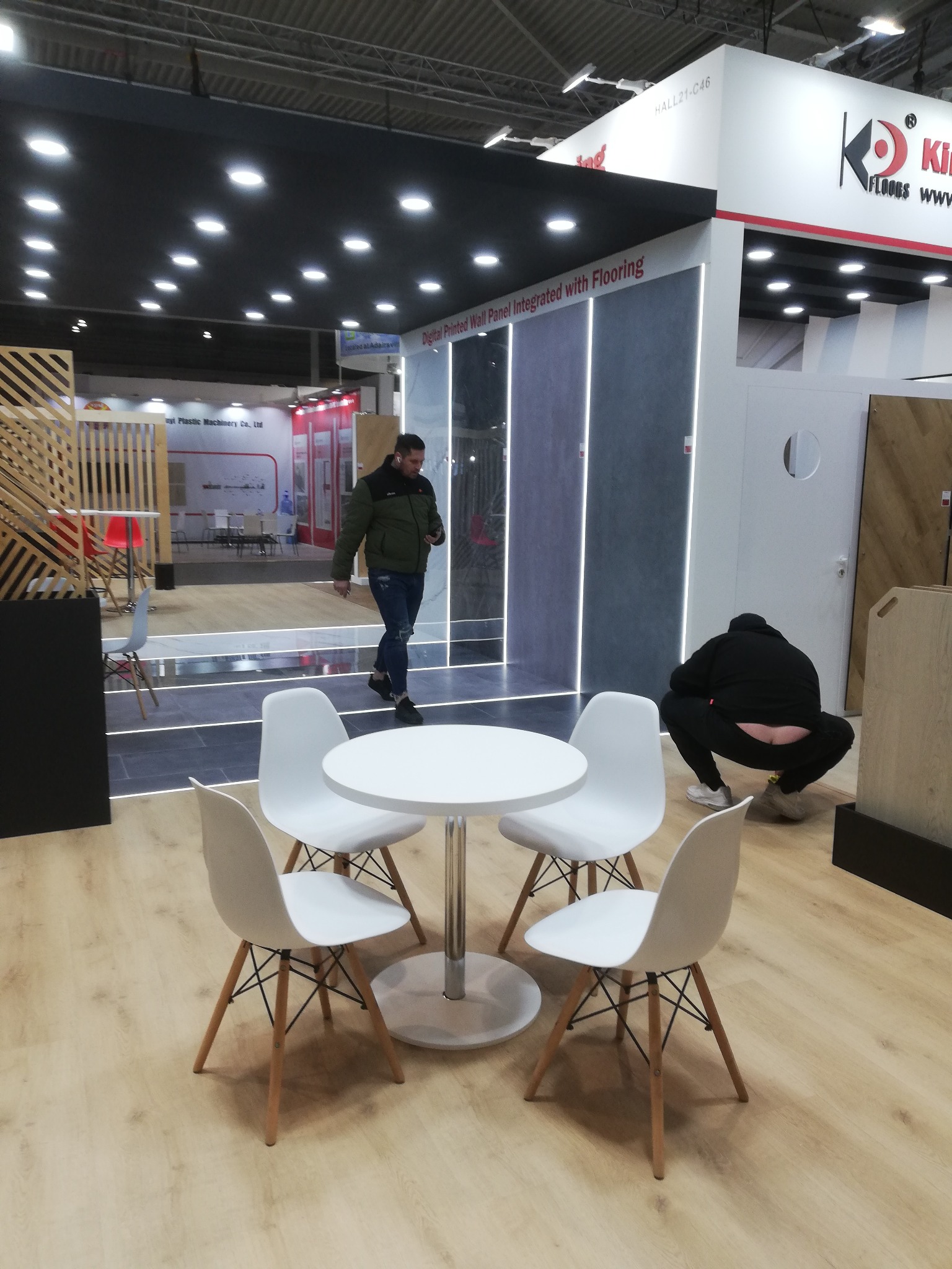 Stoisko targowe z panelami ściennymi i podłogą, oświetlone LED, z białym stolikiem i krzesłami. Osoba przechodzi obok, a inna kuca z tyłu. Widoczny fragment logo firmy.