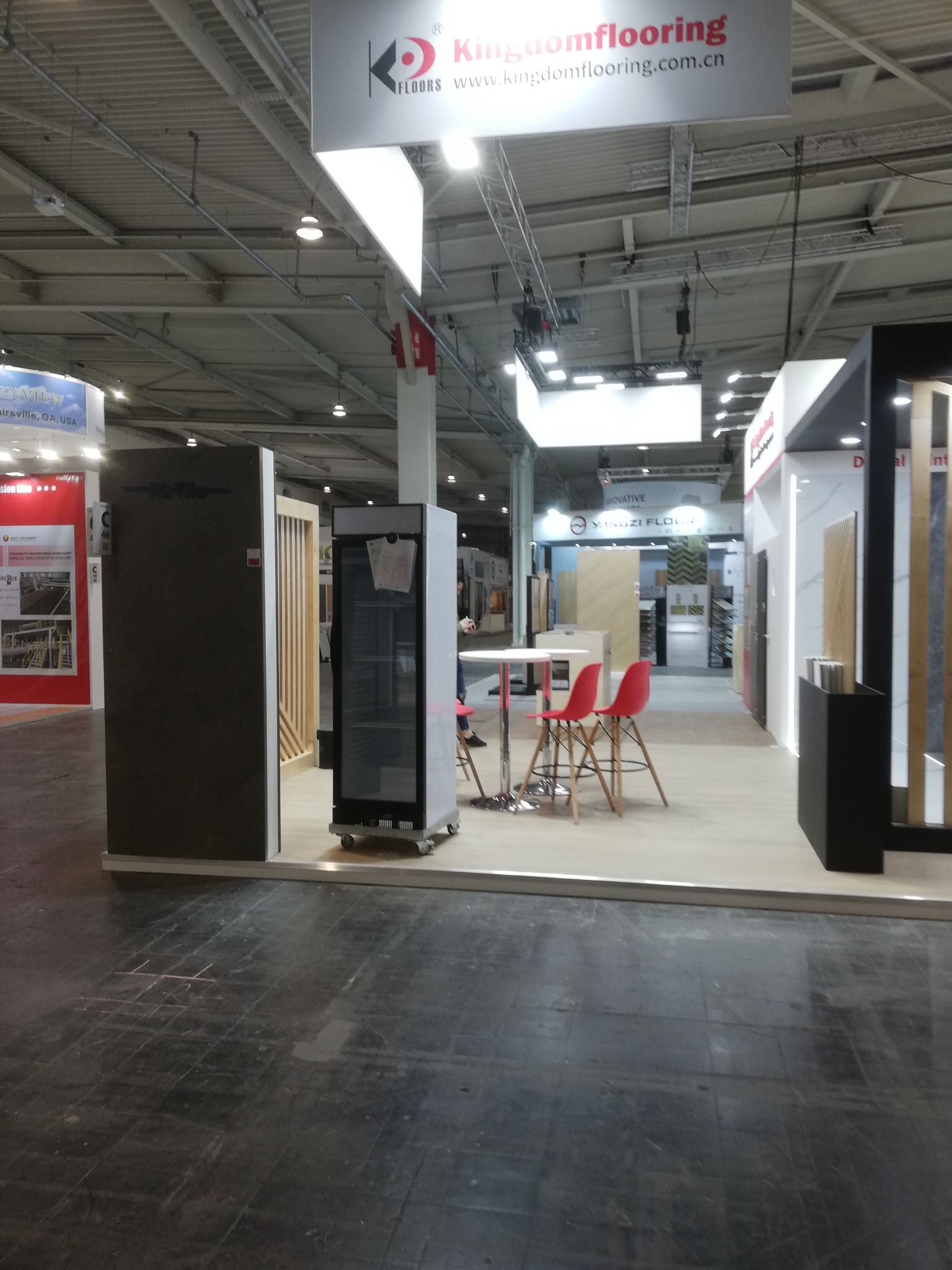 Stoisko targowe Kingdomflooring z ekspozycją paneli podłogowych, stolikiem i krzesłami. Widoczne logo firmy i oświetlenie hali.