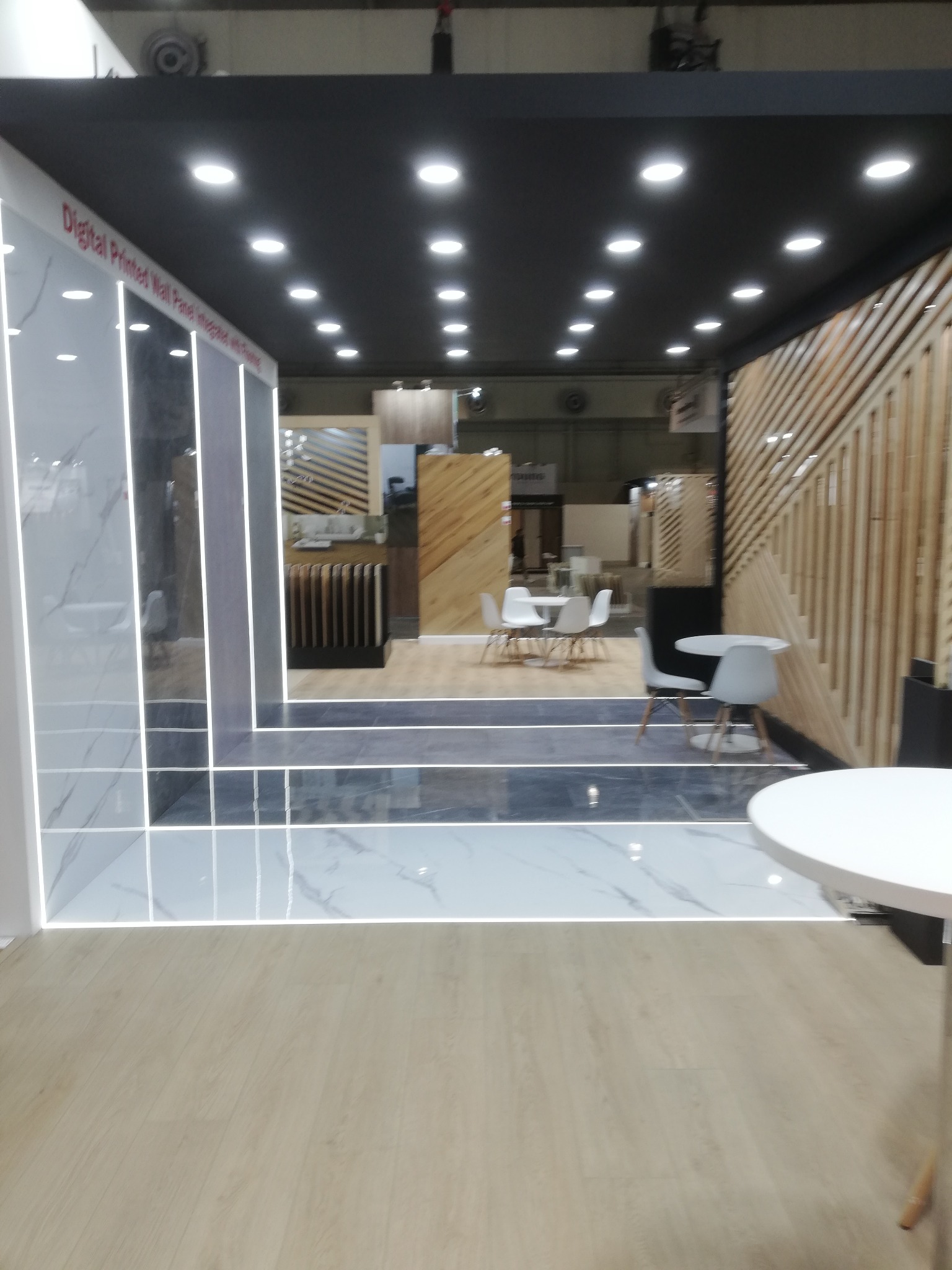 Wnętrze ekspozycji z panelami ściennymi imitującymi marmur, oświetleniem LED i drewnianymi elementami dekoracyjnymi. Widoczne stoły i krzesła w tle.