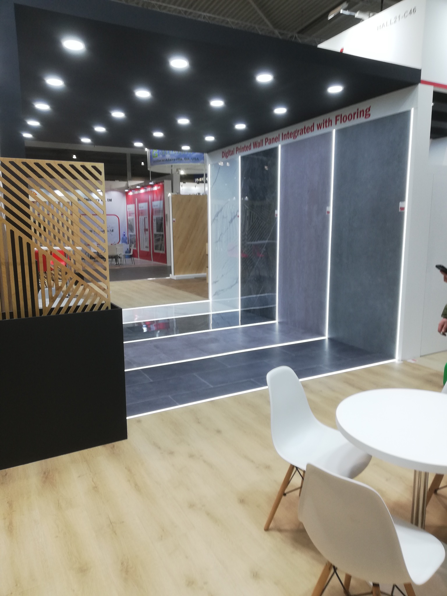 Ekspozycja paneli ściennych i podłogowych na targach. Widoczne różne wzory i kolory, oświetlenie LED, białe krzesła i stolik. Całość tworzy nowoczesną aranżację.