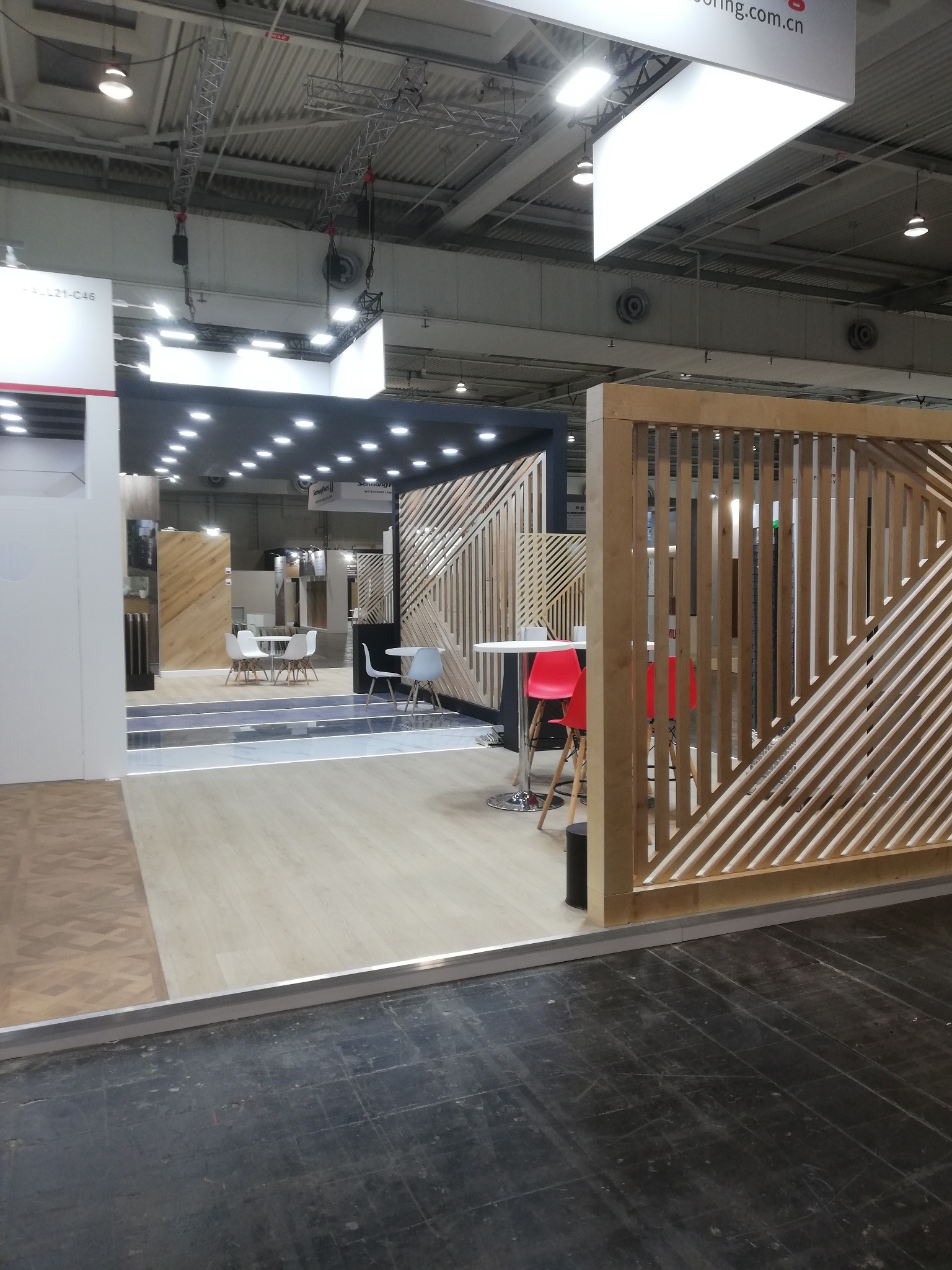 Nowoczesne stoisko targowe z drewnianą ażurową ścianką, jasną podłogą i minimalistycznymi meblami. Widoczne oświetlenie sufitowe i elementy konstrukcyjne hali.