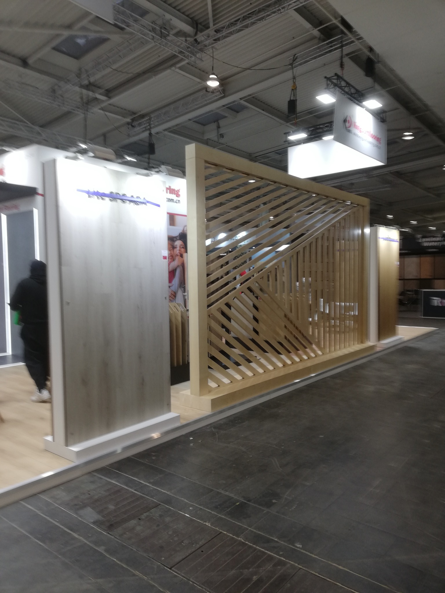 Stoisko targowe z drewnianą ażurową ścianką działową i panelami podłogowymi w odcieniach szarości i beżu. Widoczny fragment osoby i logo firmy na górze.