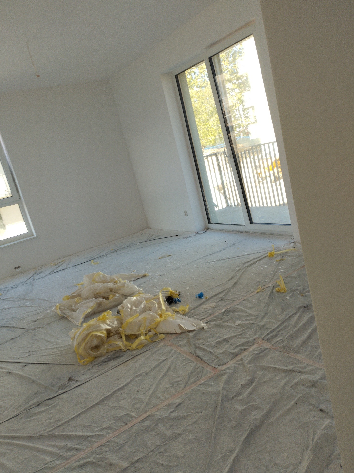 Białe ściany w pokoju z oknem balkonowym, podłoga zabezpieczona folią malarską, widoczne resztki taśmy malarskiej. Gotowe do malowania.