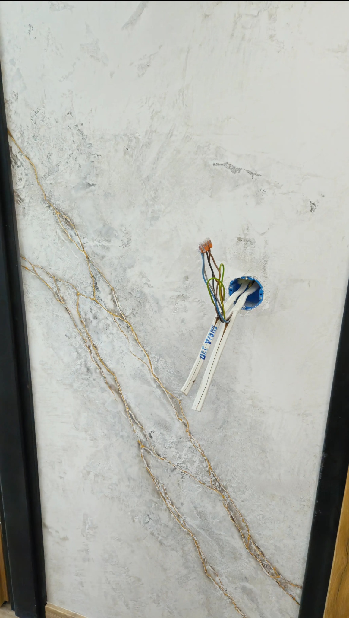 Dekoracyjna ściana z efektem marmuru i złotymi żyłkami, widoczna puszka elektryczna z przewodami. Detal wykończenia wnętrza z oryginalnym wzorem na ścianie.