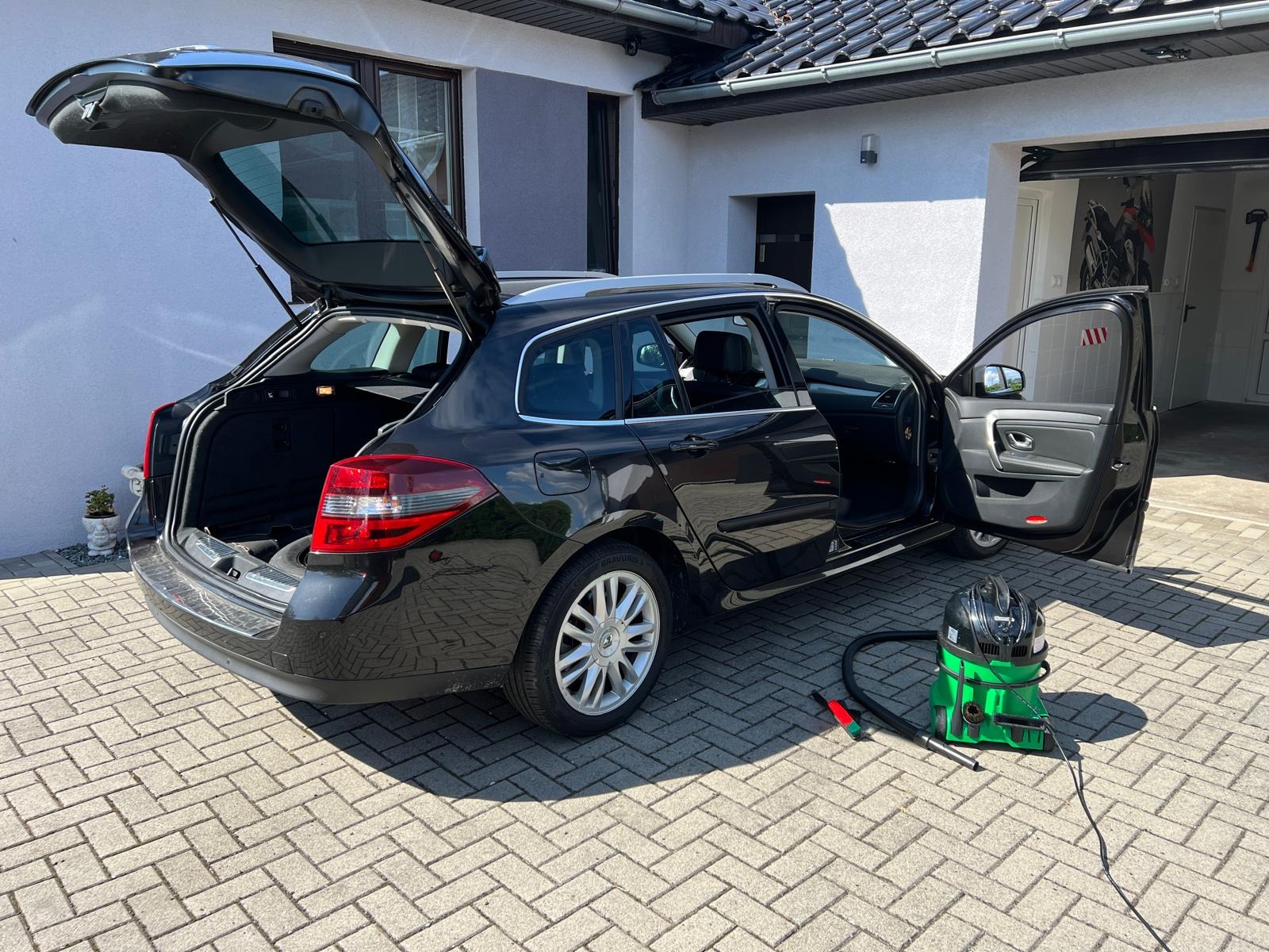Czarny samochód kombi z otwartym bagażnikiem i drzwiami, obok stoi zielony odkurzacz piorący, sugerujący czyszczenie wnętrza pojazdu na zewnątrz.