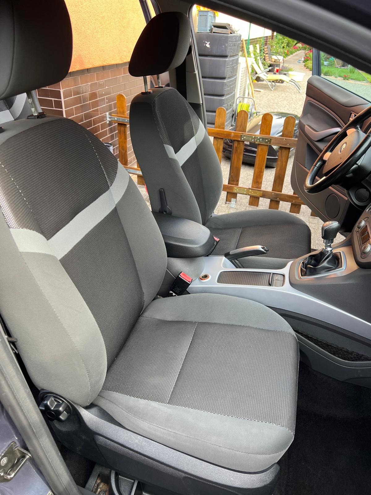 Szare fotele samochodowe z jasnymi wstawkami, widok z wnętrza auta, deska rozdzielcza, otwarte drzwi i fragment ogrodzenia w tle.