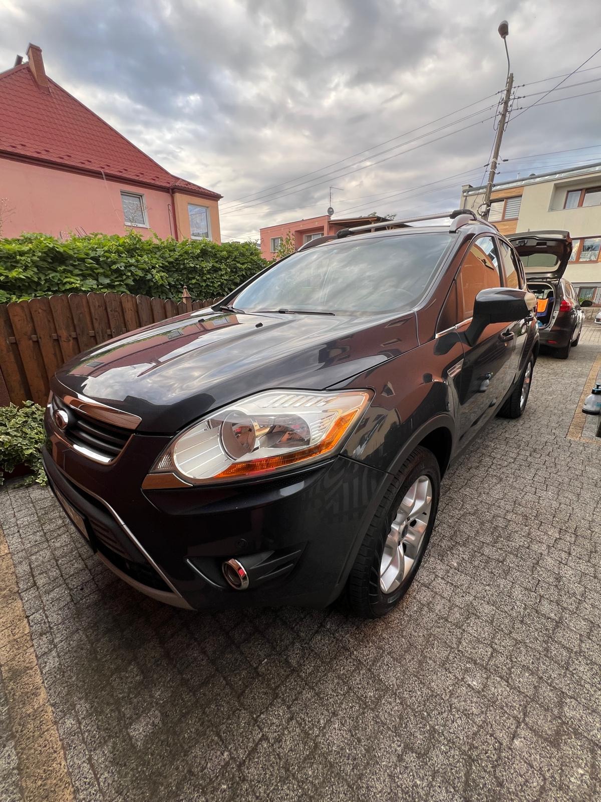 Ciemnoszary Ford Kuga na brukowanym podjeździe, z otwartym bagażnikiem drugiego auta w tle i domami w oddali, pochmurne niebo nad głową.