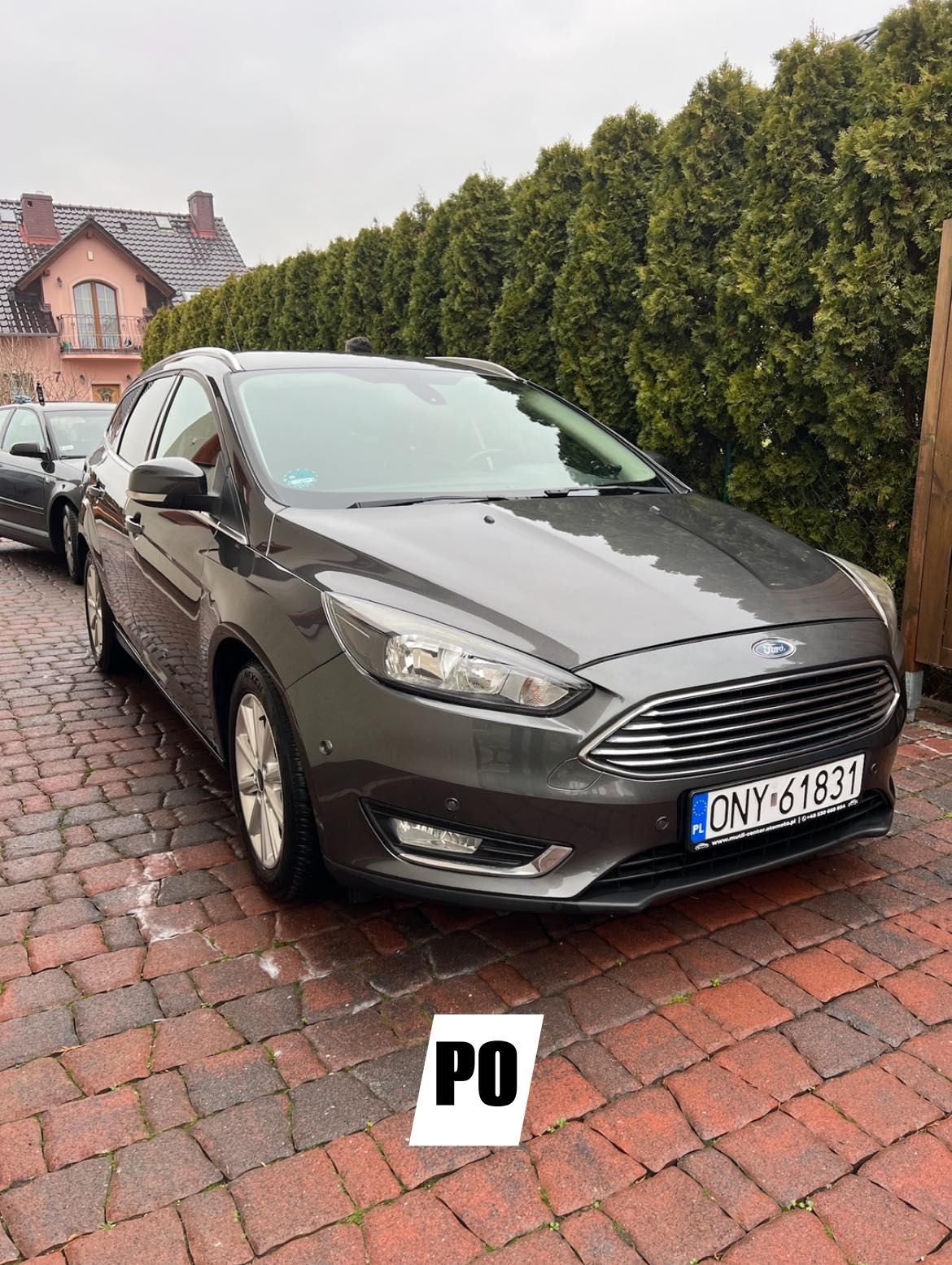 Szary Ford Focus po czyszczeniu, zaparkowany na czerwonej kostce brukowej przed zielonym żywopłotem. Widoczna tablica rejestracyjna i fragment domu w tle.
