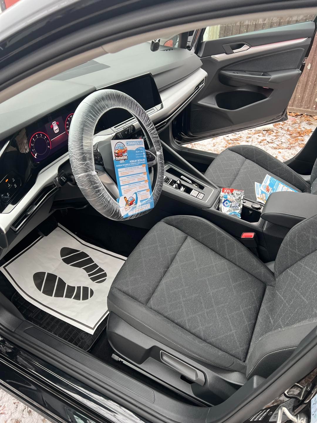 Wnętrze czarnego samochodu z zabezpieczonym folią kierownicą i wyłożonymi papierowymi matami podłogowymi. Widoczne czyste, szare fotele z materiału.
