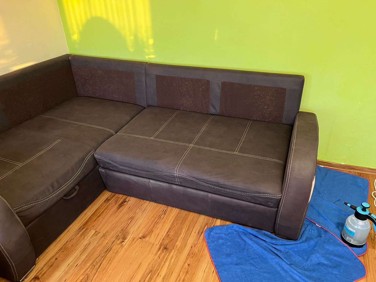 Ciemnobrązowy narożnik z funkcją spania, po praniu, na podłodze leżą niebieskie ścierki i spryskiwacz. Widoczne jasne przeszycia na tapicerce.