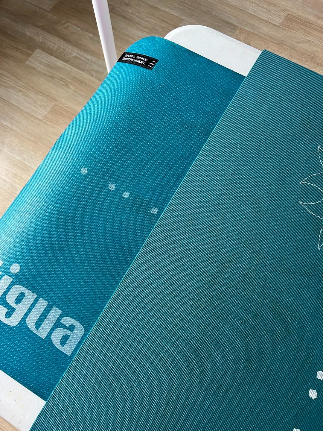 Turkusowa mata do ćwiczeń z białym napisem i wzorem kwiatowym, leżąca na białym stoliku. Widoczne detale tekstury i logo.