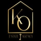 Eleganckie złote logo KO Estate Agency na czarnym tle. Sylwetka domu wkomponowana w litery, klucz zamiast litery 'I' w 'ESTATE AGENCY'. Minimalizm, prestiż.