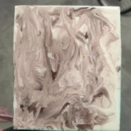 Kwadratowa próbka posadzki żywicznej w odcieniach kawy z białymi żyłkami, trzymana w dłoni na tle podłogi i roweru. Unikatowy wzór.