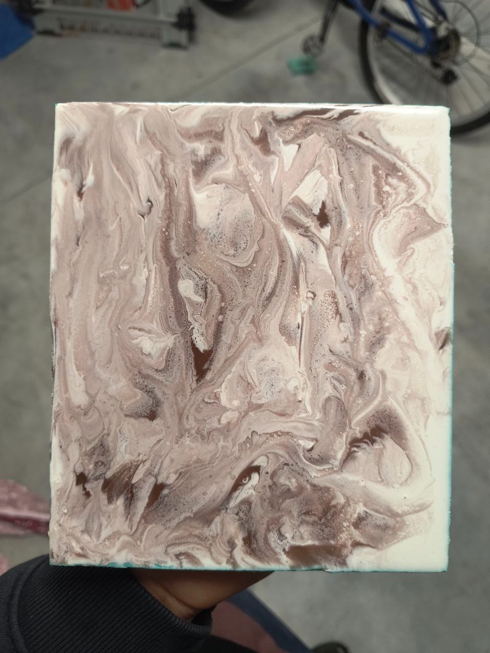 Kwadratowa próbka posadzki żywicznej w odcieniach kawy z białymi żyłkami, trzymana w dłoni na tle podłogi i roweru. Unikatowy wzór.