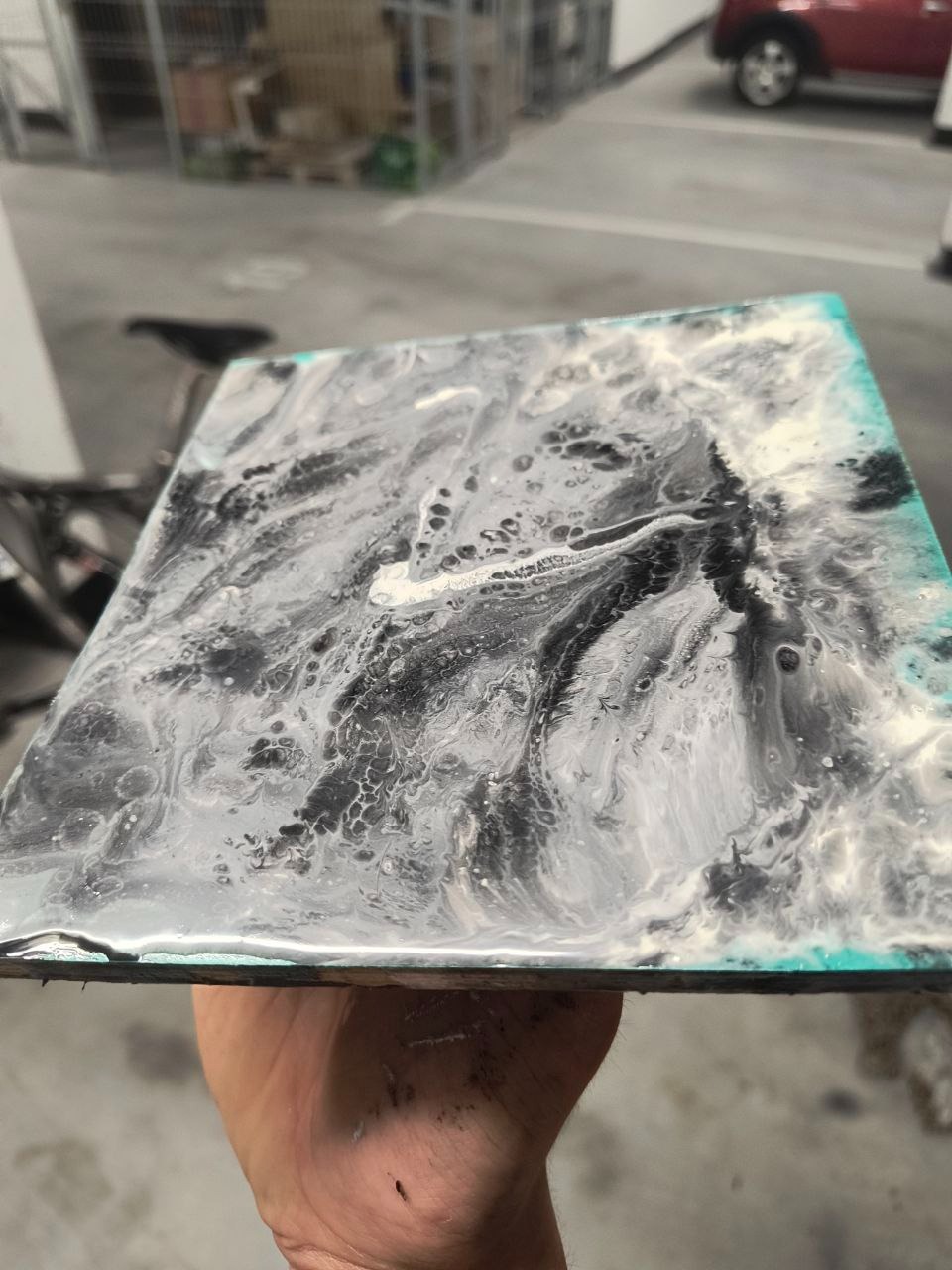 Błyszcząca, czarno-biała posadzka żywiczna, wzór marmurkowy, trzymana w dłoni. Widoczny turkusowy brzeg i struktura wylewki. Tło rozmyte, z elementami parkingu.