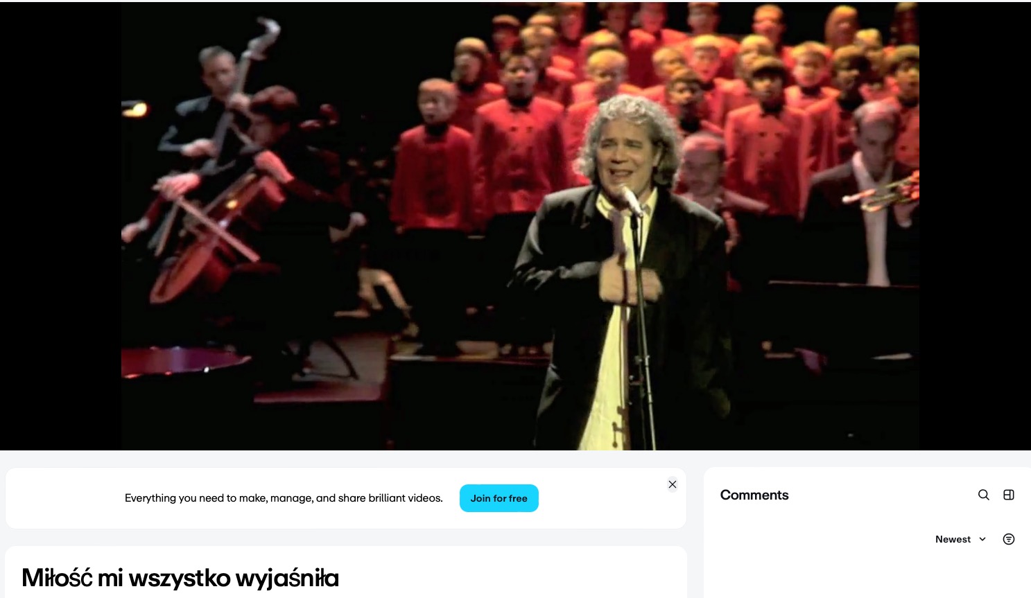 Występ chóru dziecięcego z orkiestrą i solistą. Wokalista z mikrofonem na pierwszym planie, w tle chór w czerwonych strojach i muzycy z instrumentami smyczkowymi.