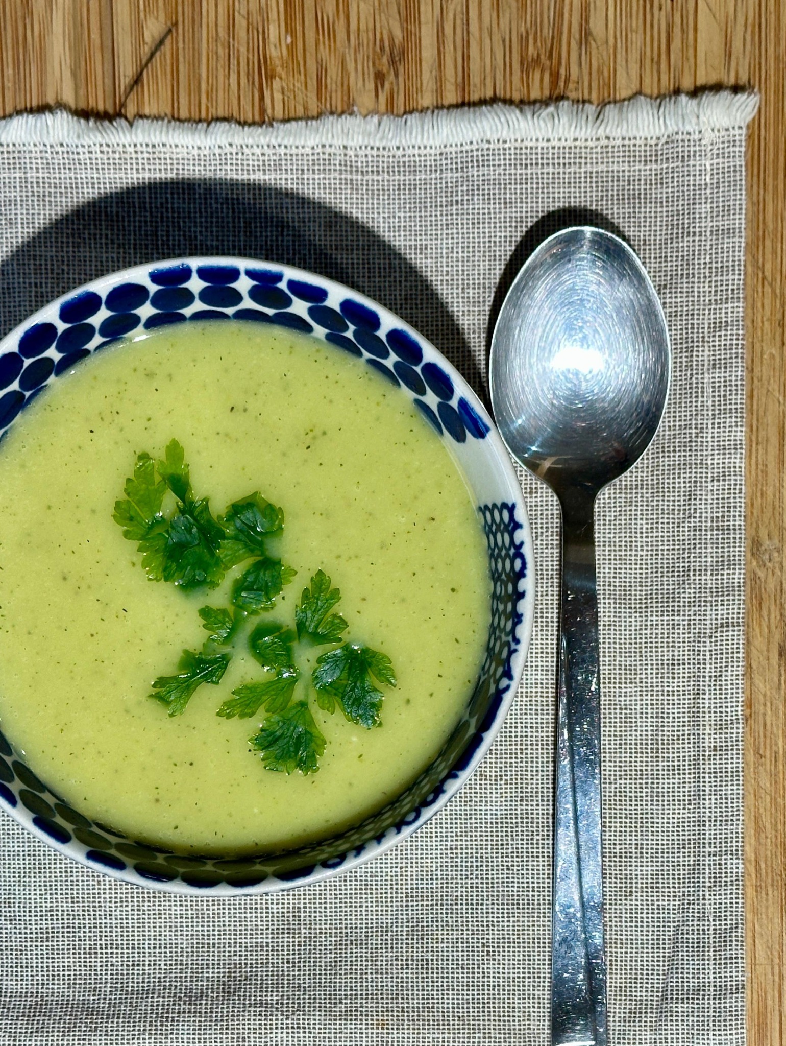 Krem z zielonych warzyw w miseczce z niebieskim wzorem, dekorowany świeżą pietruszką, obok leży łyżka na lnianej ściereczce na drewnianym stole.