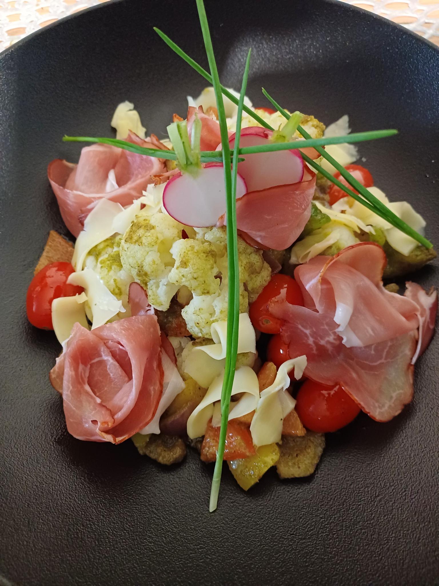 Wykwintna sałatka na czarnym talerzu: różyczki z szynki parmeńskiej, rzodkiewki, pomidorki cherry i zielony szczypiorek. Elegancki catering na specjalne okazje.