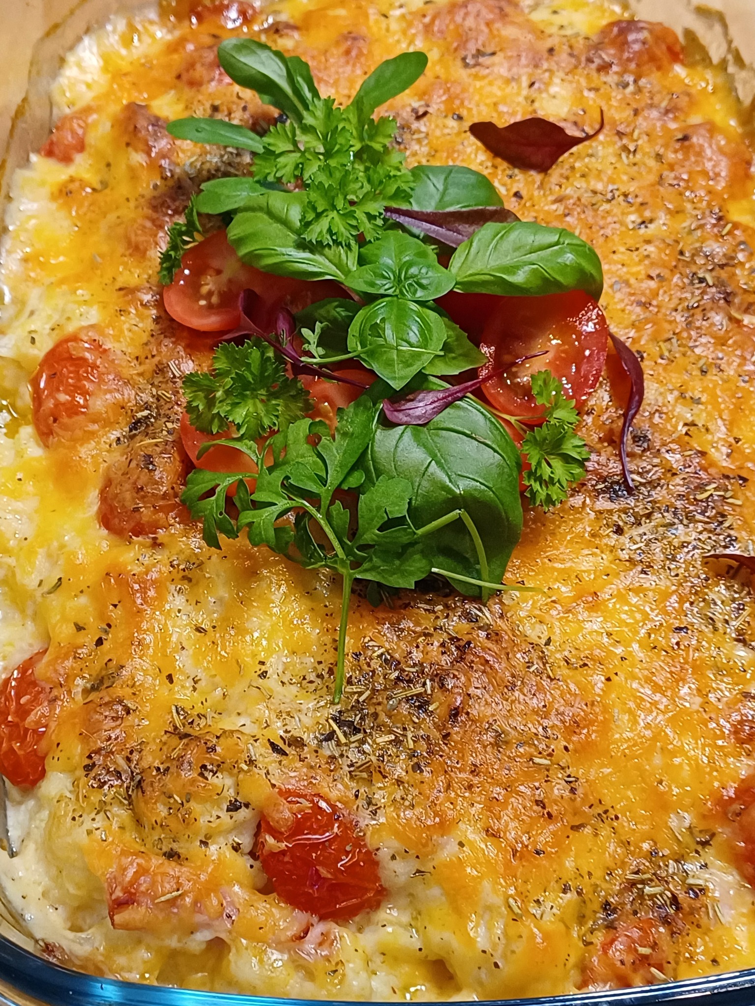 Zapiekanka z pomidorkami cherry i ziołami, podana w szklanym naczyniu żaroodpornym, z dekoracją z bazylii i pietruszki. Danie gotowe do spożycia.