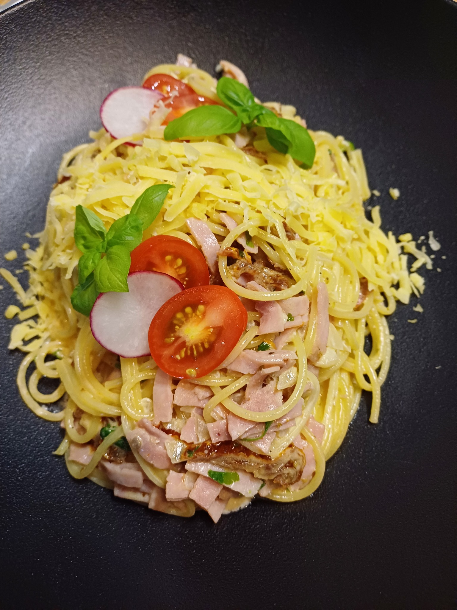 Apetyczne spaghetti z szynką, serem i świeżymi warzywami na czarnym talerzu, udekorowane bazylią. Kulinarna kompozycja gotowa do serwowania.