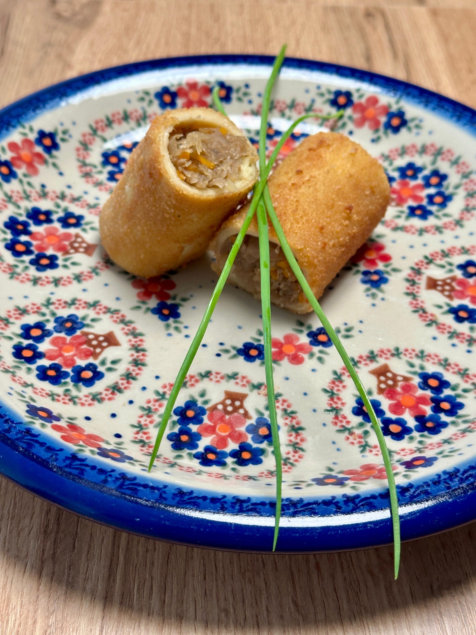 Krokiety z kapustą i mięsem, przekrojone na pół, udekorowane szczypiorkiem, ułożone na talerzu w stylu Bolesławiec na drewnianym stole.