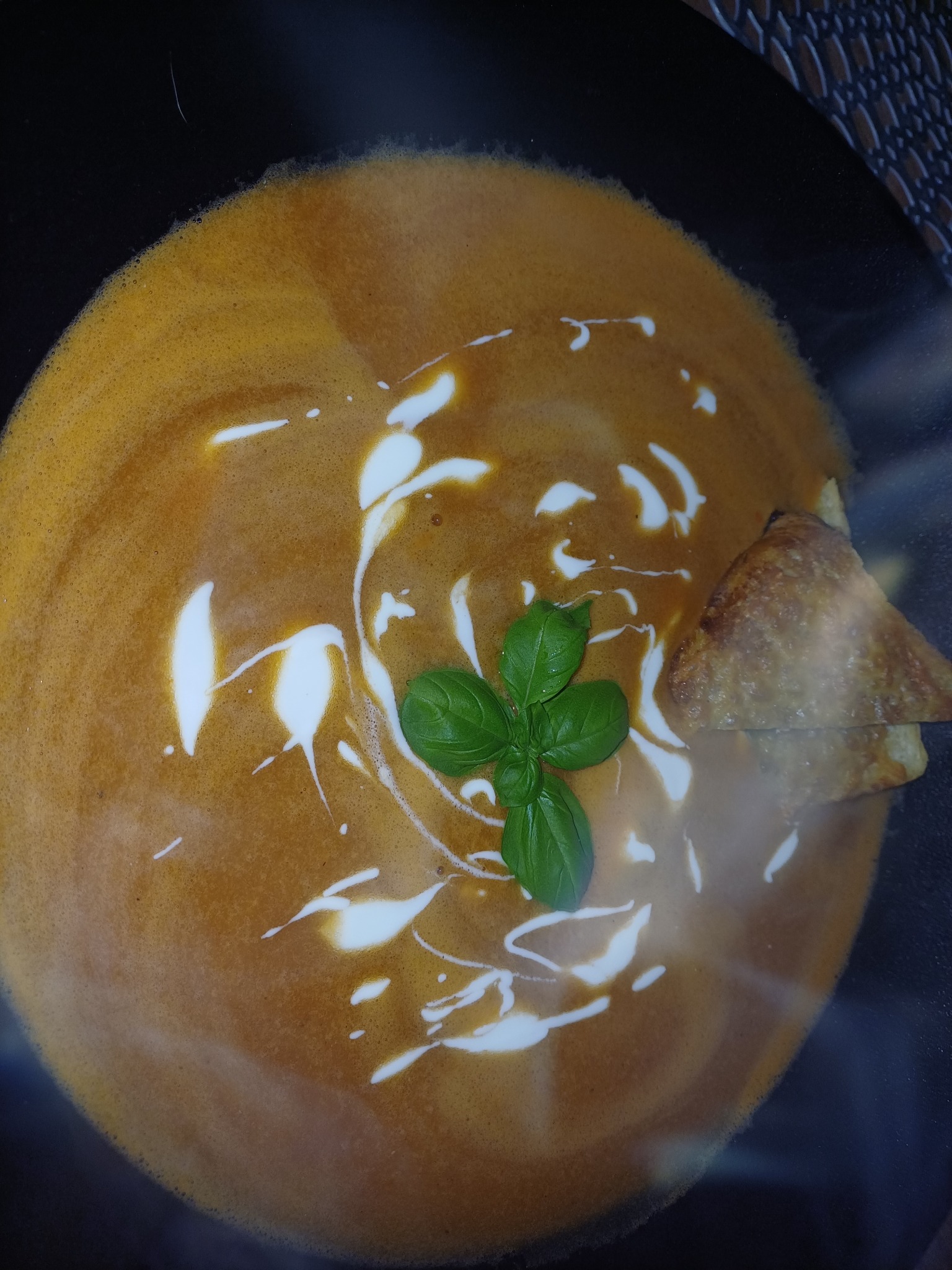 Krem pomidorowy w czarnym talerzu, zdobiony kleksem śmietany i listkiem bazylii, z dodatkiem trójkątnej grzanki. Elegancki sposób podania zupy.