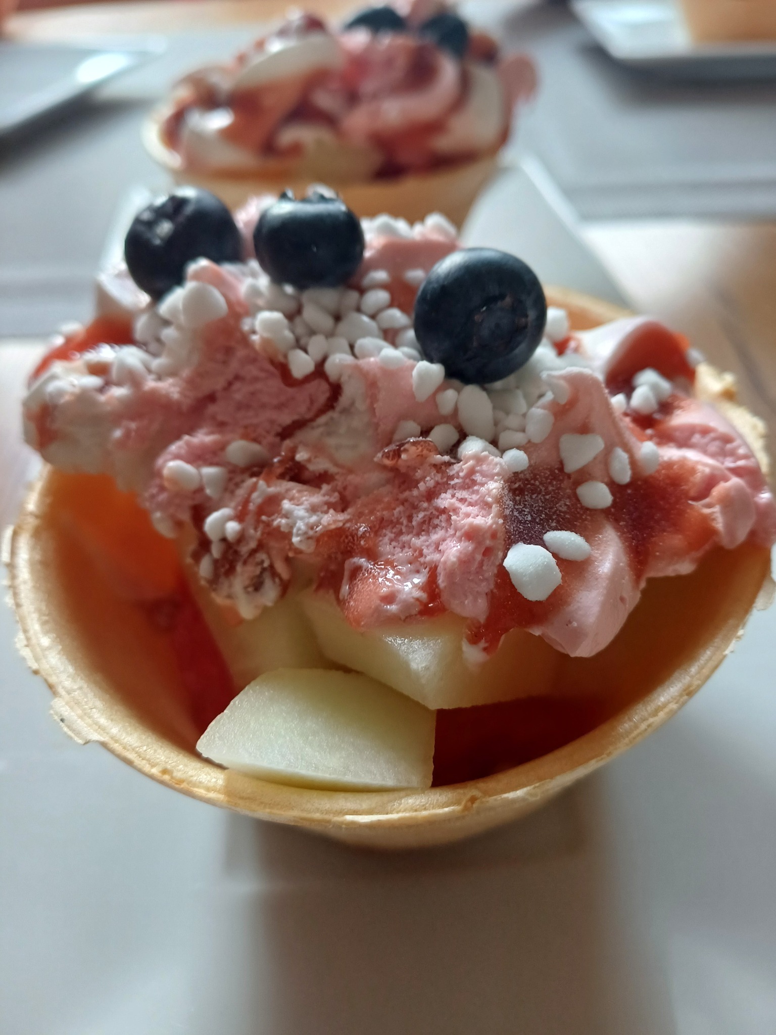 Deser w waflowym koszyczku z owocami, bitą śmietaną, polewą i borówkami, udekorowany drobnymi, białymi kuleczkami. Widoczne dwa desery, ustawione na białym tle.