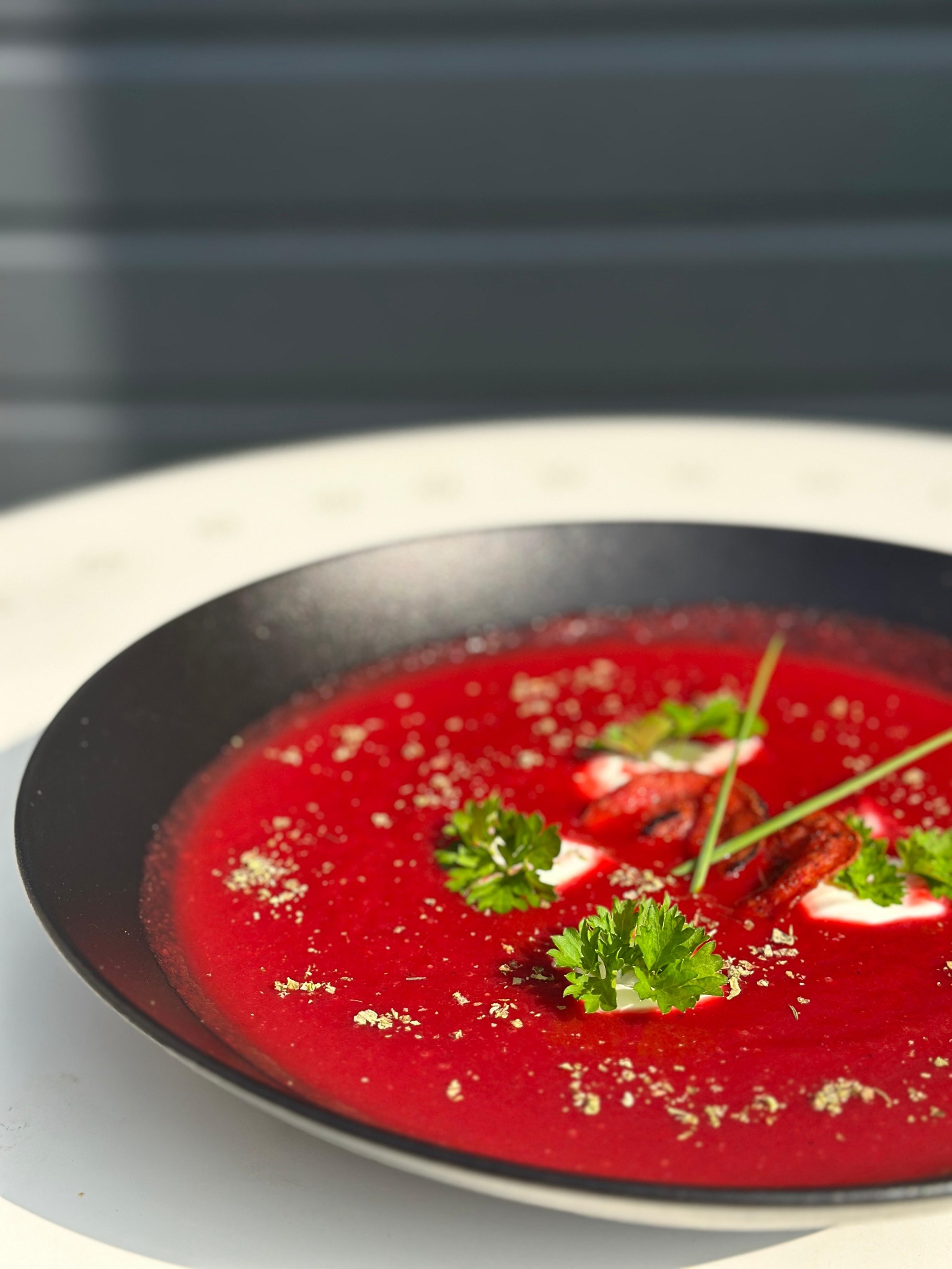 Krem z buraków w czarnym talerzu, udekorowany natką pietruszki, suszonymi pomidorami i ziołami, na jasnym stole, w naturalnym oświetleniu.