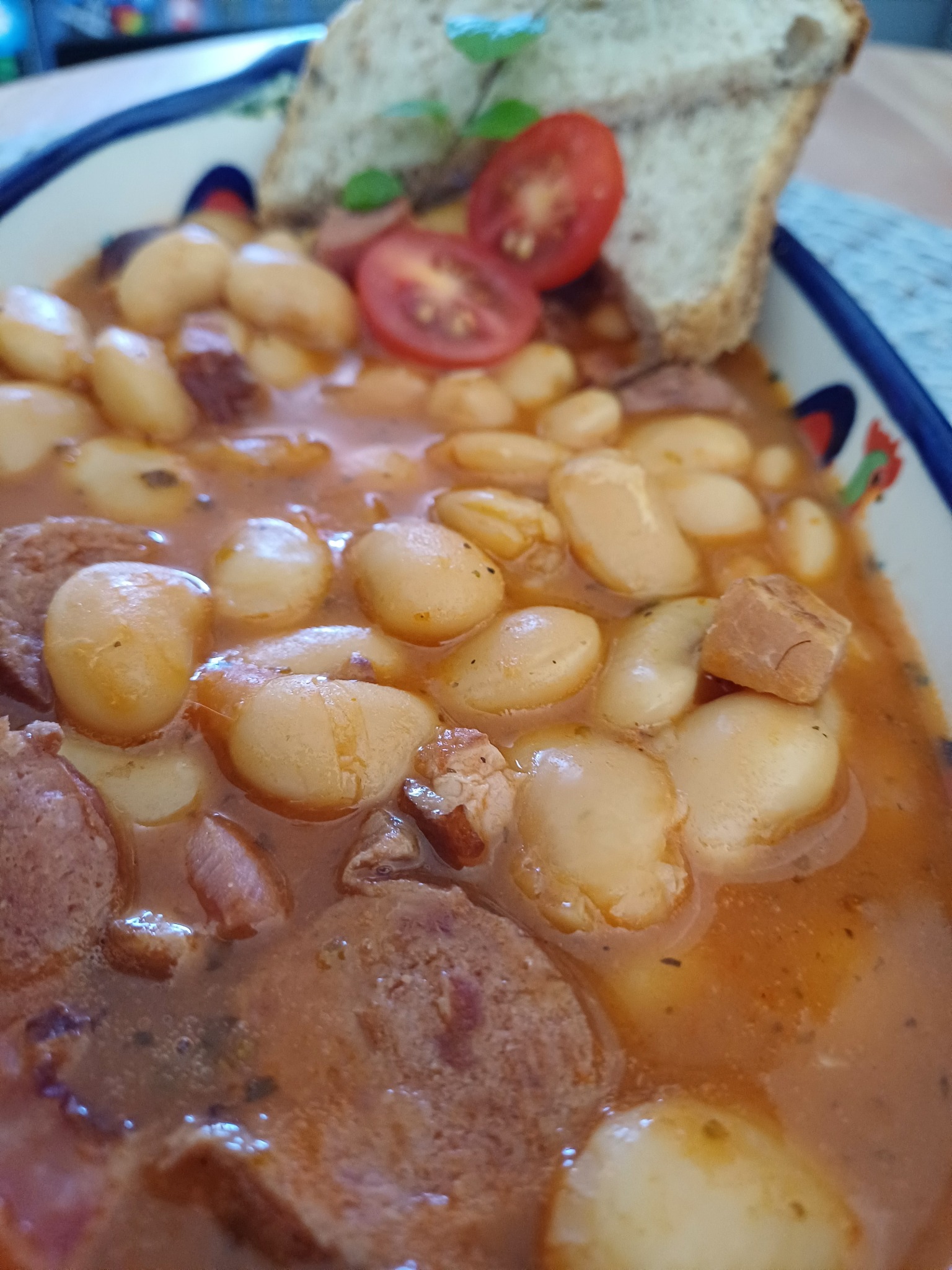 Fasolka po bretońsku w zdobionej misce, z kromką chleba, pomidorami i bazylią. Widoczne kawałki kiełbasy i boczku. Domowy obiad w zbliżeniu.