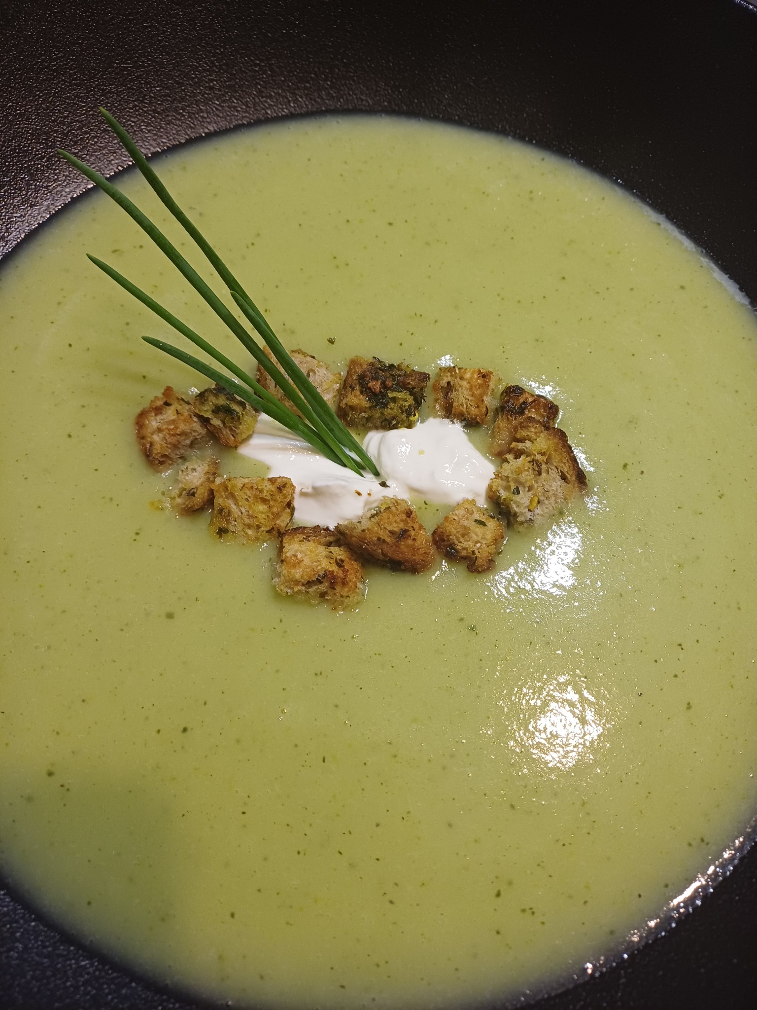 Krem z zielonych warzyw w czarnym talerzu, udekorowany grzankami, kleksem śmietany i szczypiorkiem. Eleganckie podanie, zbliżenie na detale.