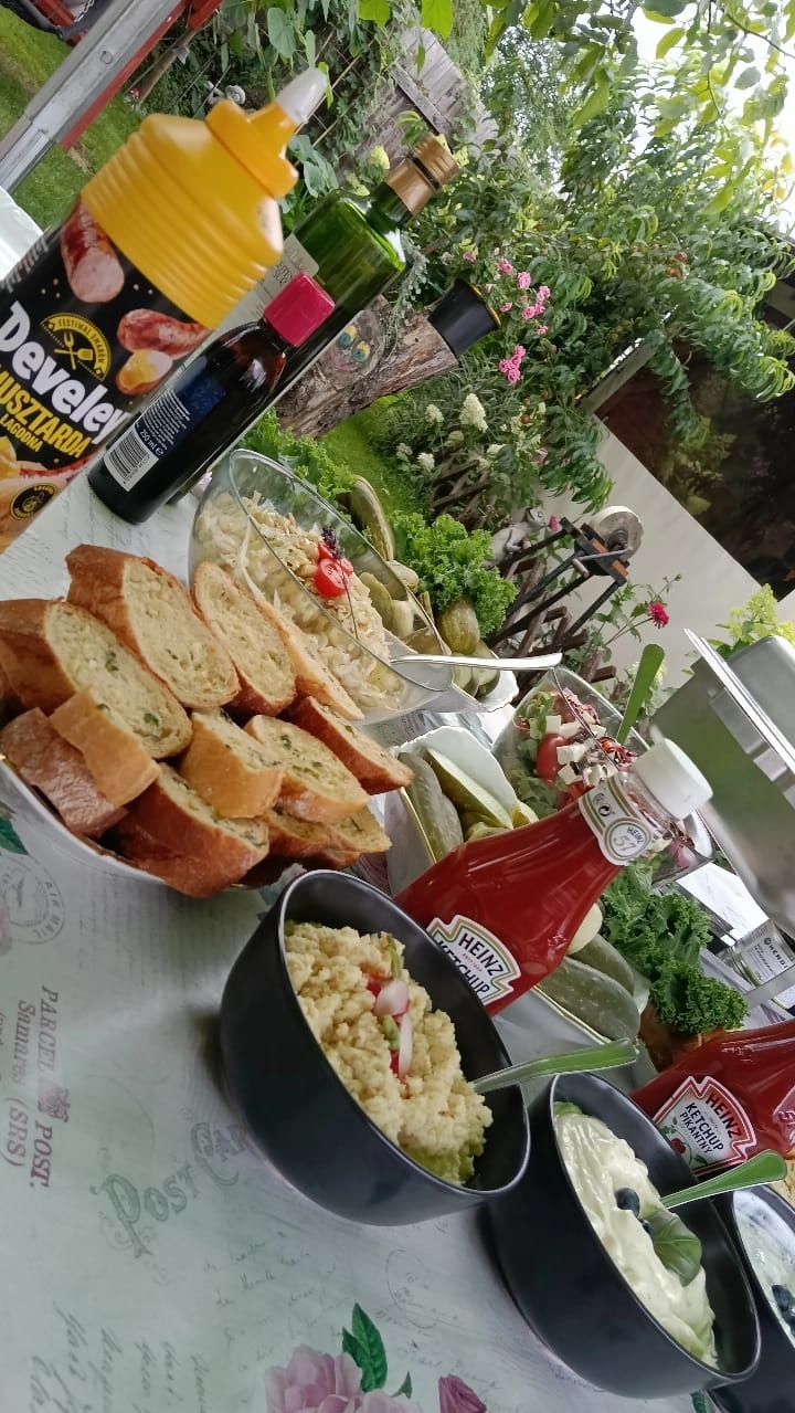Stół cateringowy z pieczywem, sałatkami, ogórkami, musztardą Develey i ketchupem Heinz w plenerowej scenerii, z zielenią w tle. Różnorodność potraw.