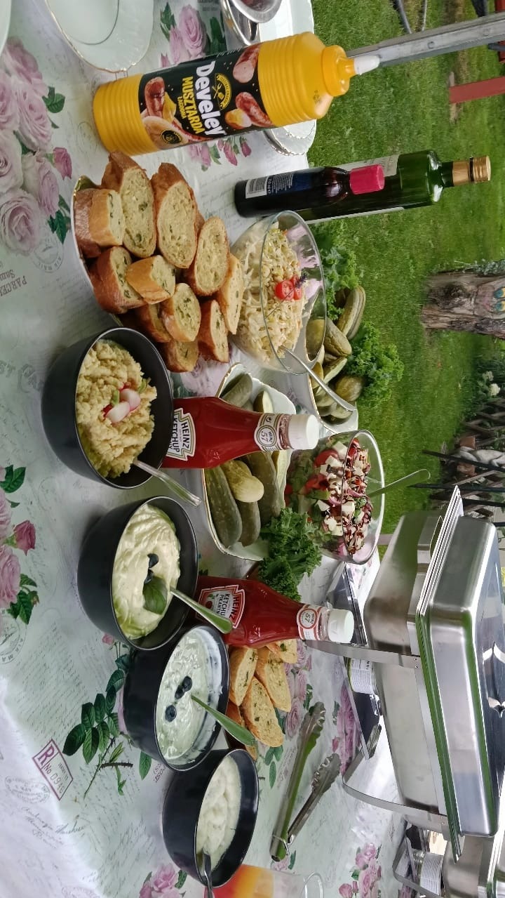 Stół cateringowy na zewnątrz: sałatki, pieczywo, sosy, musztarda Develey, ketchup Heinz. Dekoracyjny obrus w kwiaty, trawa w tle. Elementy zastawy stołowej.