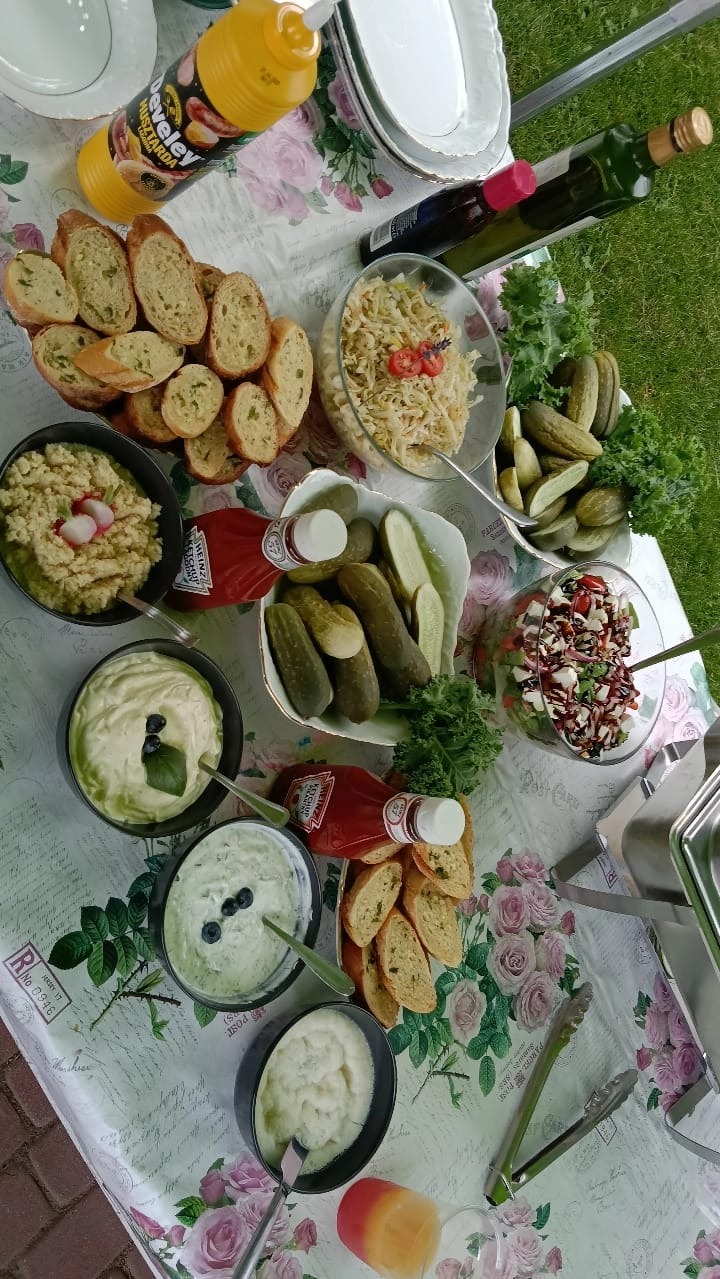 Stół cateringowy z różnorodnymi sałatkami, dipami, pieczywem i marynatami na kwiecistym obrusie, z butelkami sosów i oliwy, gotowy na przyjęcie w plenerze.
