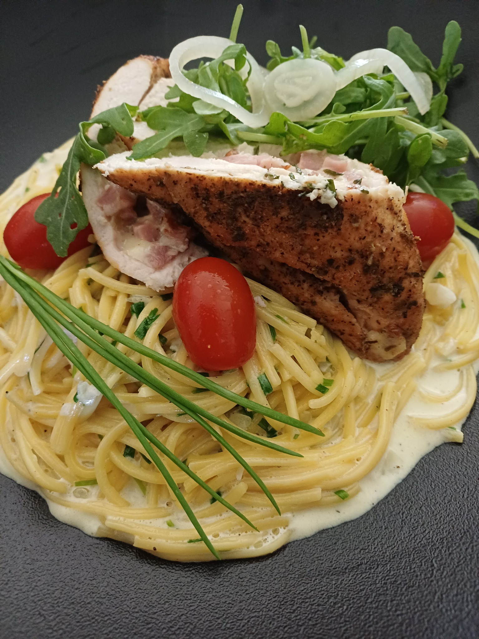 Elegancko podane spaghetti z grillowanym kurczakiem faszerowanym szynką, rukolą, pomidorkami koktajlowymi i szczypiorkiem na czarnym talerzu.