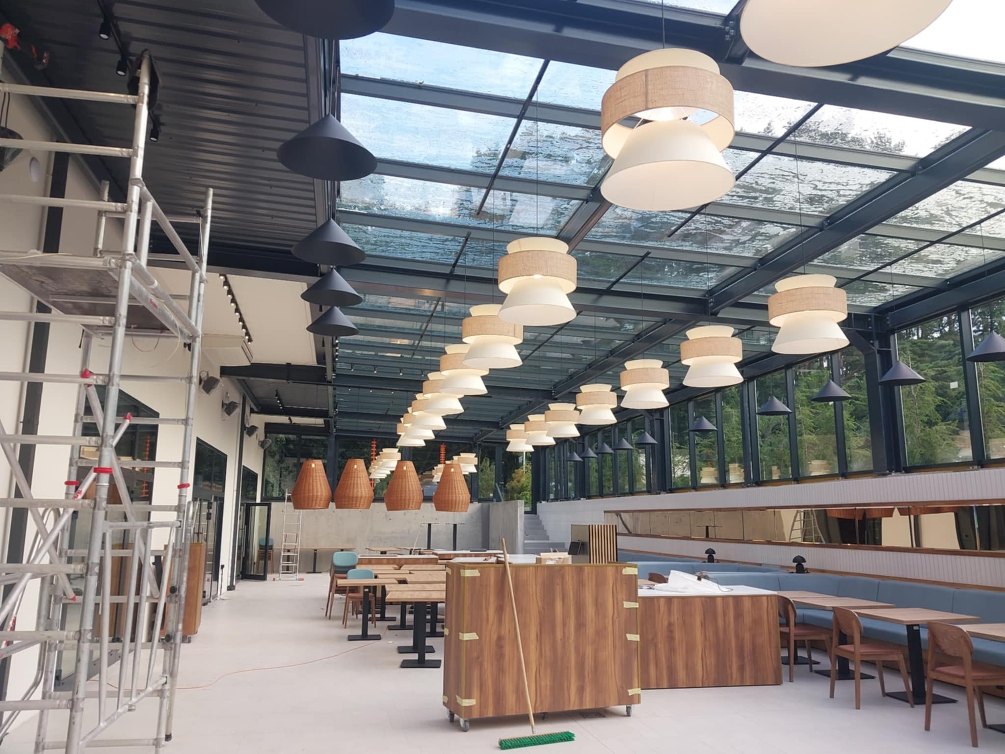 Montaż oświetlenia w restauracji z przeszklonym dachem. Widoczne lampy wiszące, rusztowanie i elementy wyposażenia wnętrza.