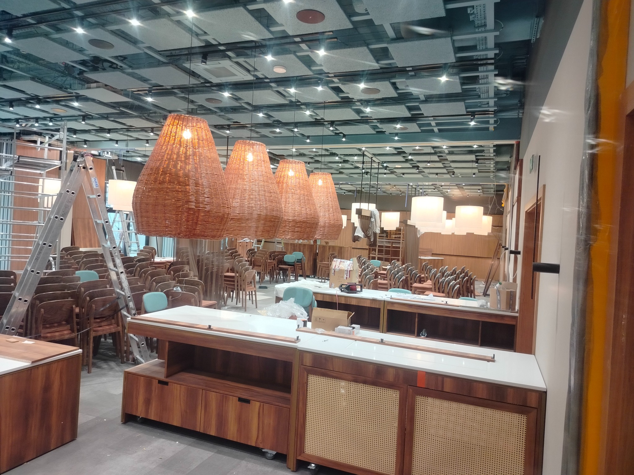 Wnętrze lokalu w trakcie wykańczania: wiszące, plecione lampy nad ladą, krzesła, drabina malarska, sufit z oświetleniem. Widoczne prace wykończeniowe.