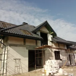 Dom w stanie surowym otwartym z czarną dachówką i widocznymi rynnami, otoczony rusztowaniami i materiałami budowlanymi, pod błękitnym niebem.