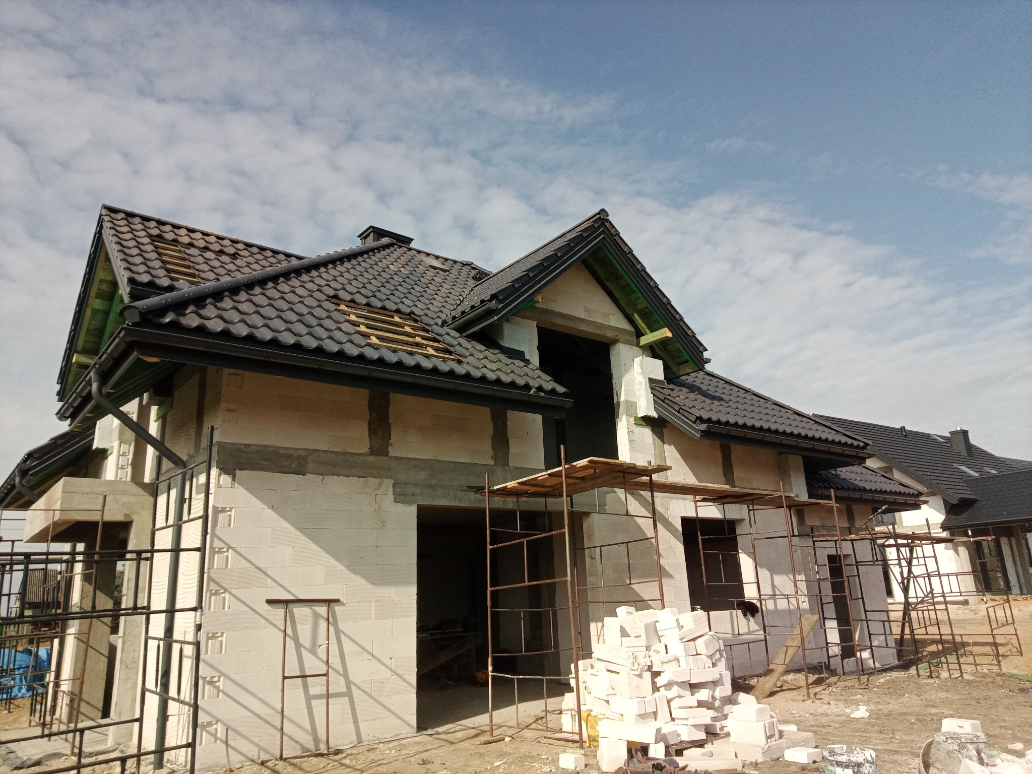Dom w stanie surowym otwartym z czarną dachówką i widocznymi rynnami, otoczony rusztowaniami i materiałami budowlanymi, pod błękitnym niebem.