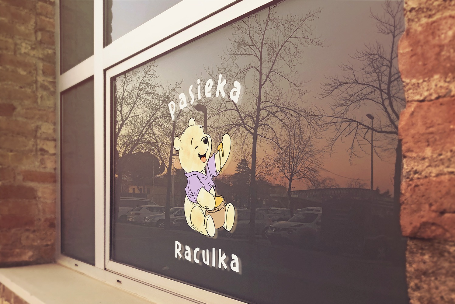 Grafika z wizerunkiem Kubusia Puchatka i napisem 'Pasieka Raculka' umieszczona na szybie okiennej lokalu, widok z zewnątrz, z ulicą i drzewami w tle.