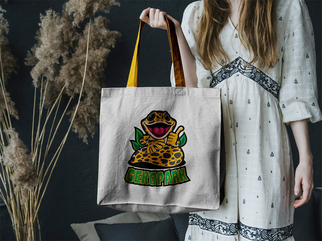 Biała torba z nadrukiem wesołego gekona z podniesionym kciukiem, trzymana przez kobietę w jasnej sukience. W tle ciemna ściana i trawa pampasowa.