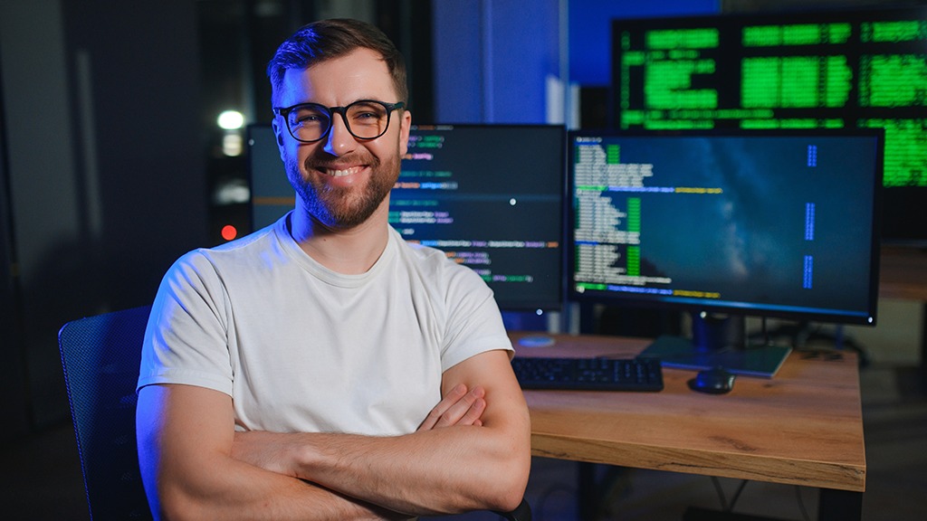 Uśmiechnięty programista w okularach, z założonymi rękami, siedzi przed biurkiem z dwoma monitorami wyświetlającymi kod. W tle widać dodatkowy ekran z zielonym kodem.