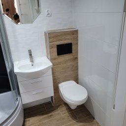 Kacper Cyranski - Nowoczesna łazienka z biało-drewnianymi płytkami, umywalką z szafką i toaletą. Lustro odbija osobę robiącą zdjęcie. Widoczny fragment kabiny prysznicowej.