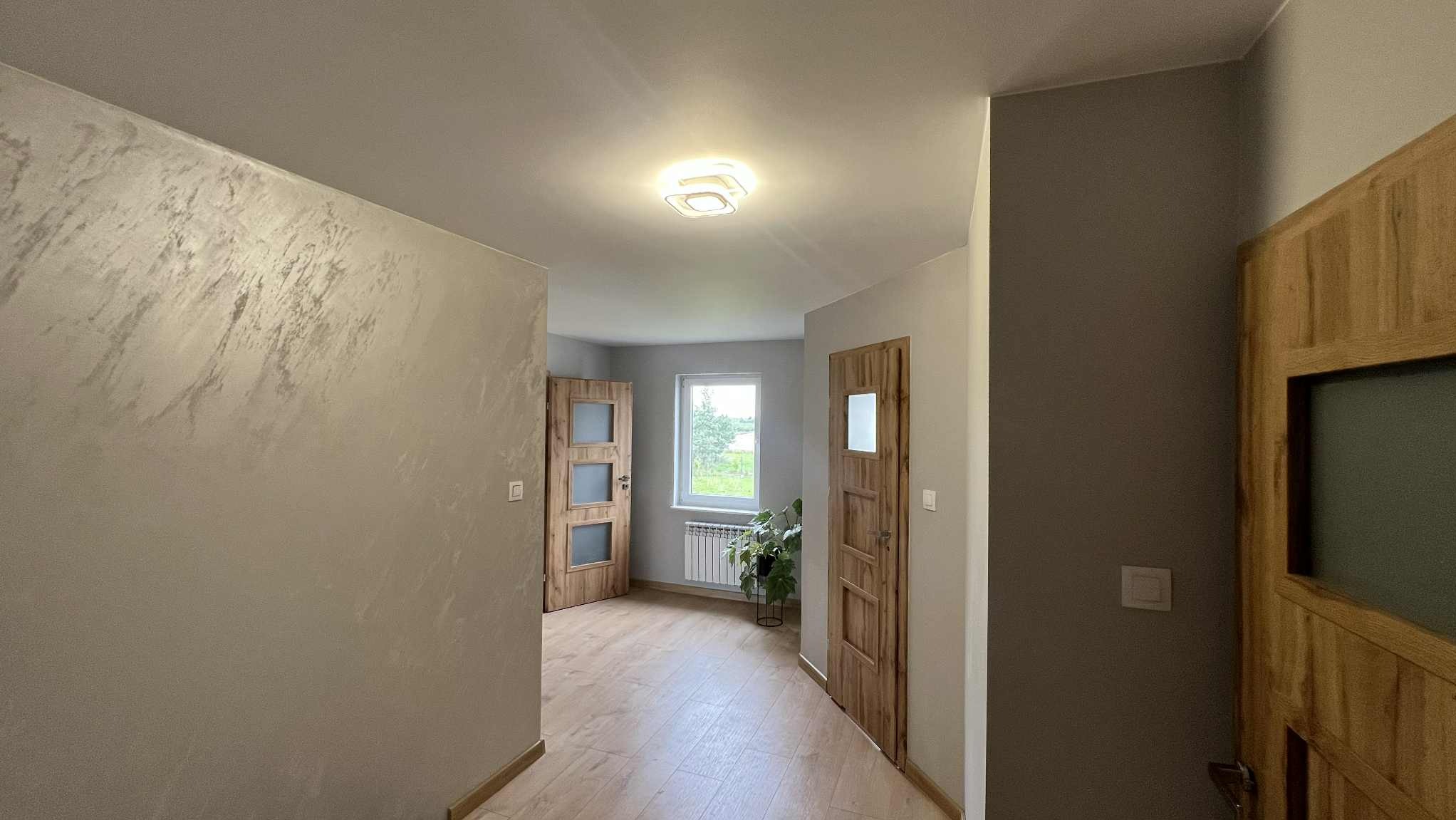 Wykończony korytarz z drewnianymi drzwiami, dekoracyjną szpachlą na ścianie i jasną podłogą. Widok na okno z rośliną. Nowoczesne oświetlenie sufitowe.