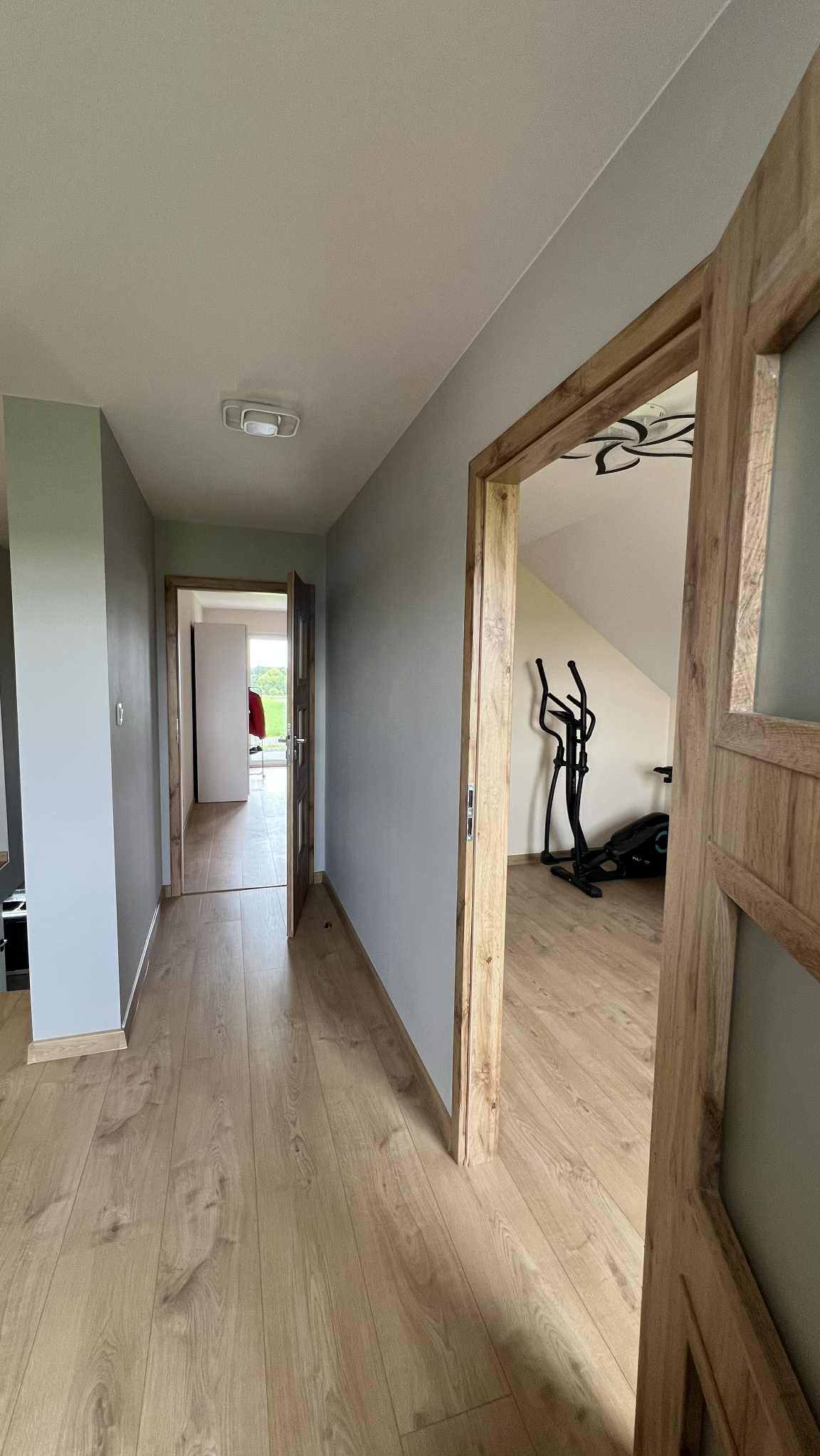 Korytarz z drewnianą podłogą i szarymi ścianami. Widoczne otwarte drzwi z drewnianą ościeżnicą, prowadzące do pokoju z sprzętem do ćwiczeń. Minimalistyczne oświetlenie sufitowe.