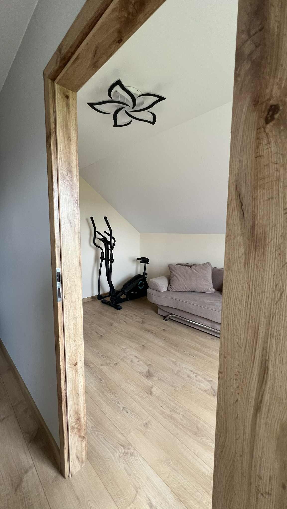 Widok przez drewnianą futrynę na poddasze z podłogą panelową, sofą, orbitrekiem i rowerem stacjonarnym. Na suficie lampa w kształcie kwiatu.