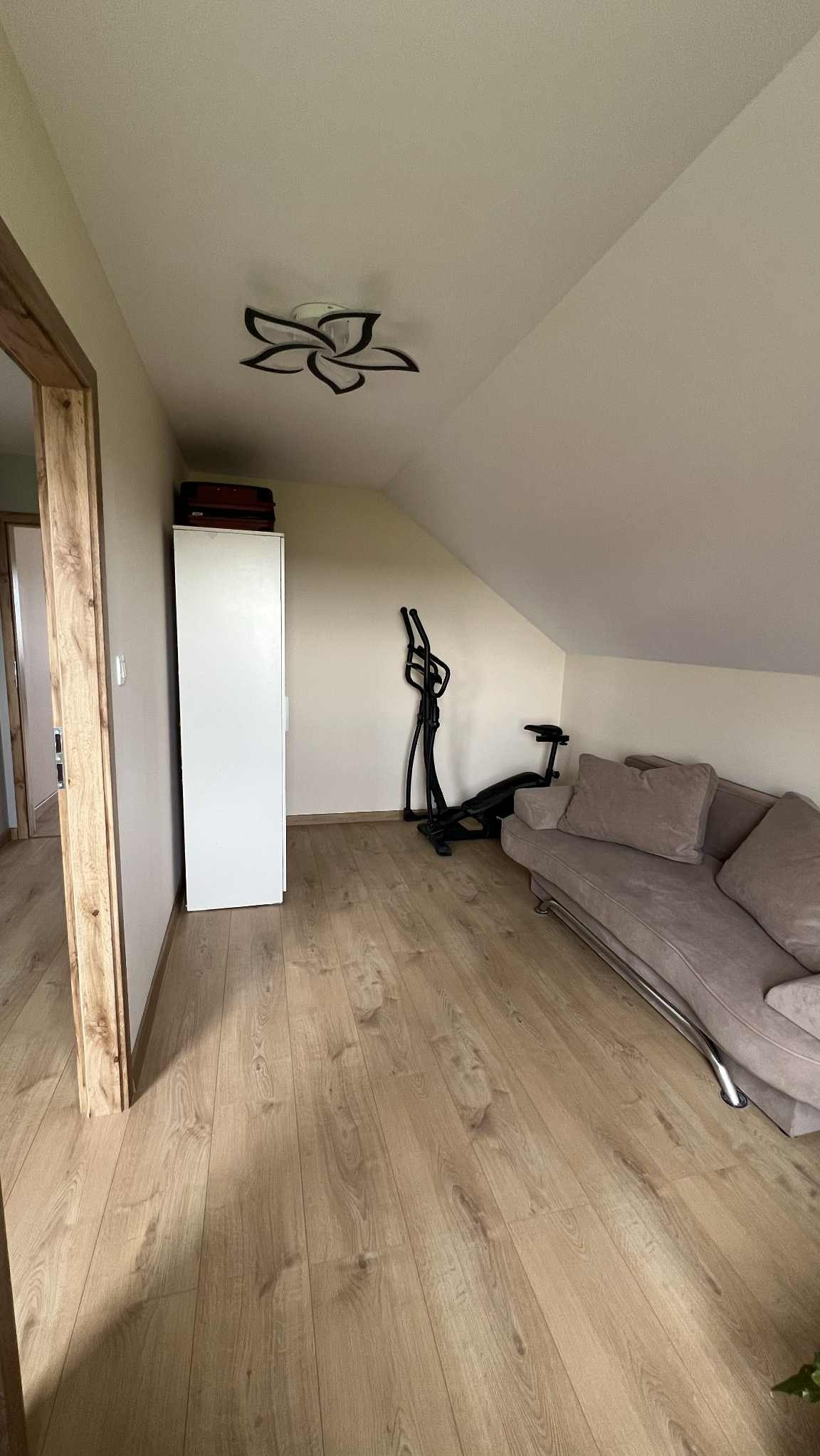 Jasne poddasze z drewnianą podłogą i skośnym sufitem. Biała szafa, sofa i sprzęt do ćwiczeń. Lampa sufitowa w kształcie kwiatu. Widok z perspektywy wejścia.