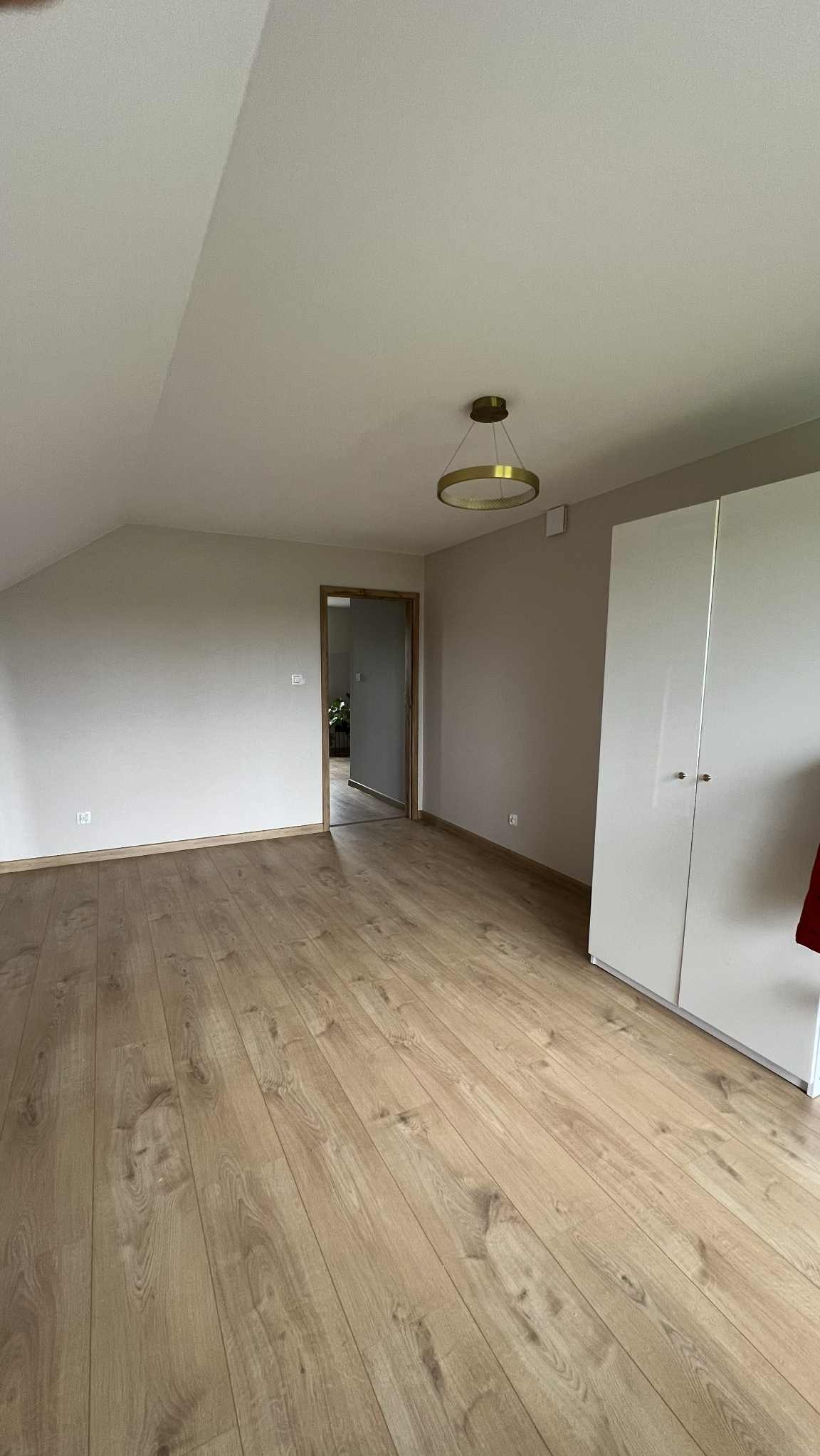 Jasne panele podłogowe w pokoju na poddaszu z pomalowanymi na beżowo ścianami. Widoczna biała szafa i drzwi do kolejnego pomieszczenia. Okrągła lampa sufitowa.
