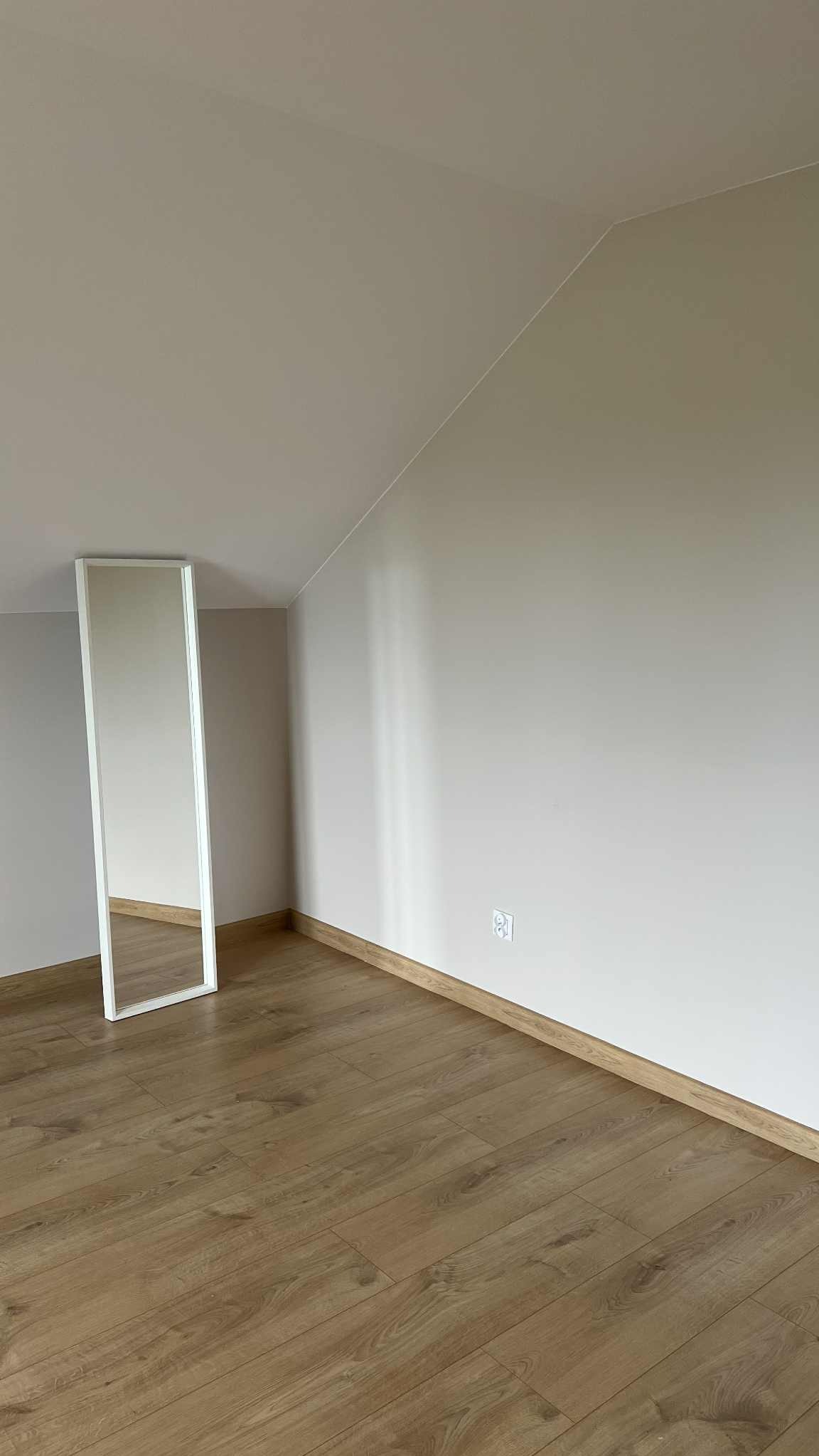 Jasne wnętrze poddasza z drewnianą podłogą i białym lustrem opartym o ścianę. Widoczne skosy sufitu i gniazdko elektryczne.