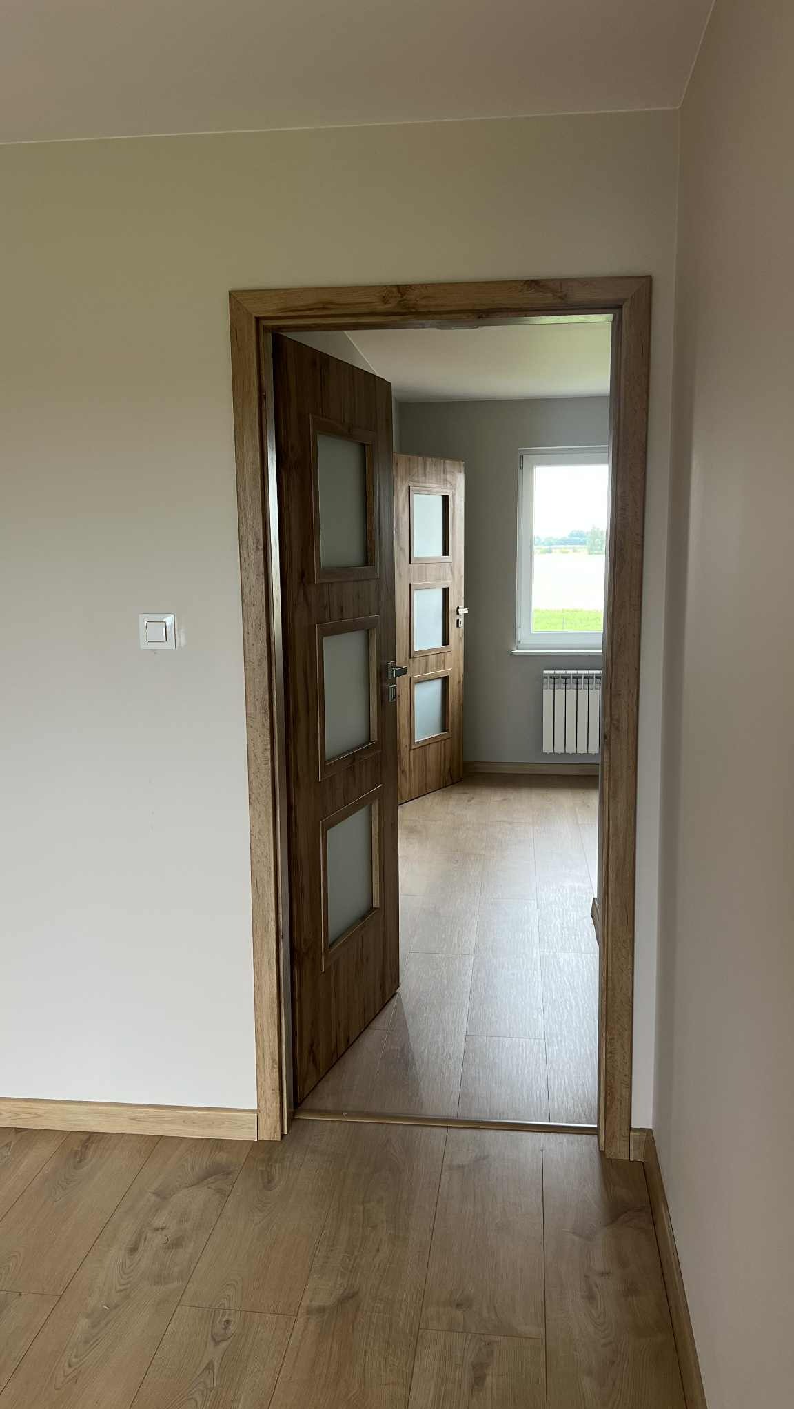 Wnętrze z drewnianymi drzwiami z szybkami, widoczne kolejne drzwi i okno z grzejnikiem. Jasne ściany i podłoga z paneli. Widok z perspektywy korytarza.
