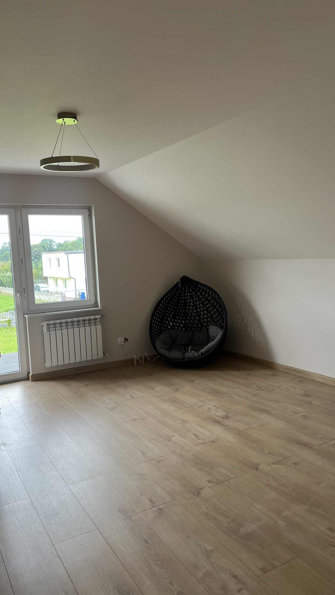 Jasne wnętrze poddasza z drewnianą podłogą, oknem i wiszącym fotelem. Ściany pomalowane na biało, nowoczesna lampa sufitowa. Widok na zieleń za oknem.